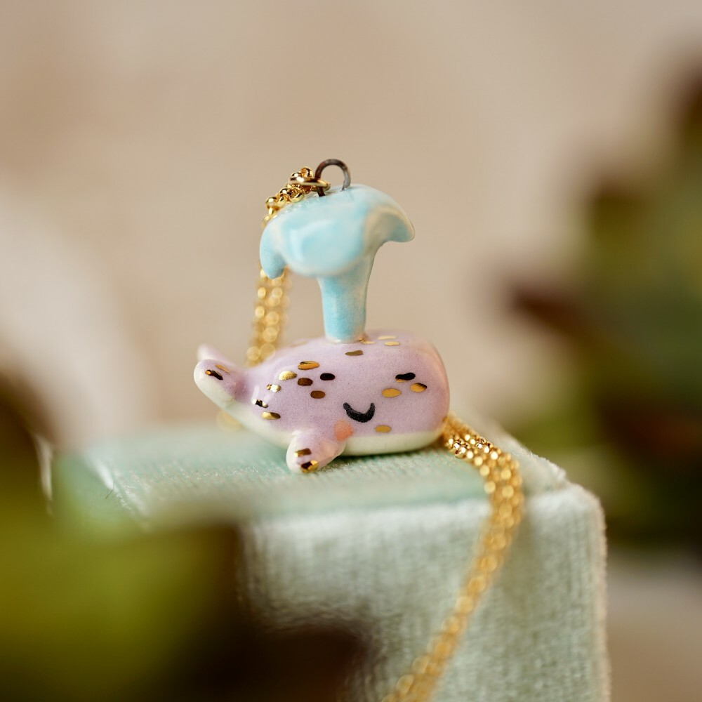 Twee pink ceramic spouting whale pendant with light blue spout and gold details attached to a gold chain.