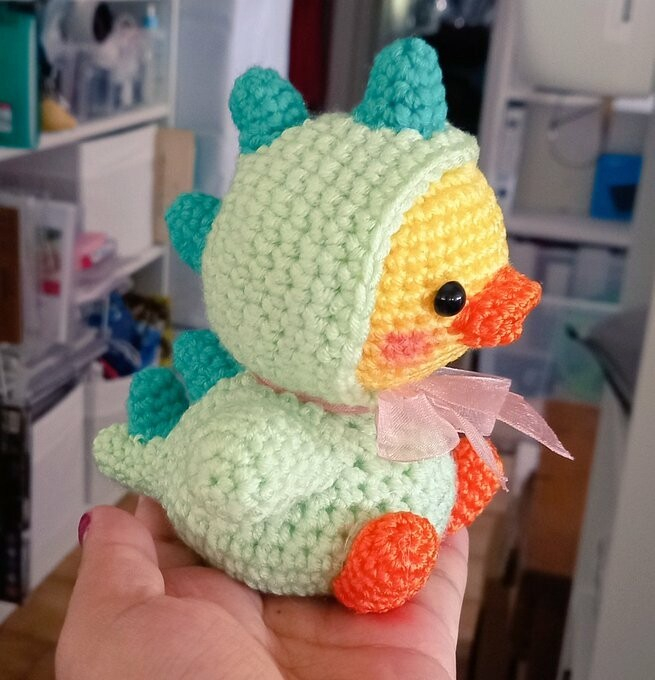 Photograph of a cute crocheted duckling wearing a pale green dinosaur costume.