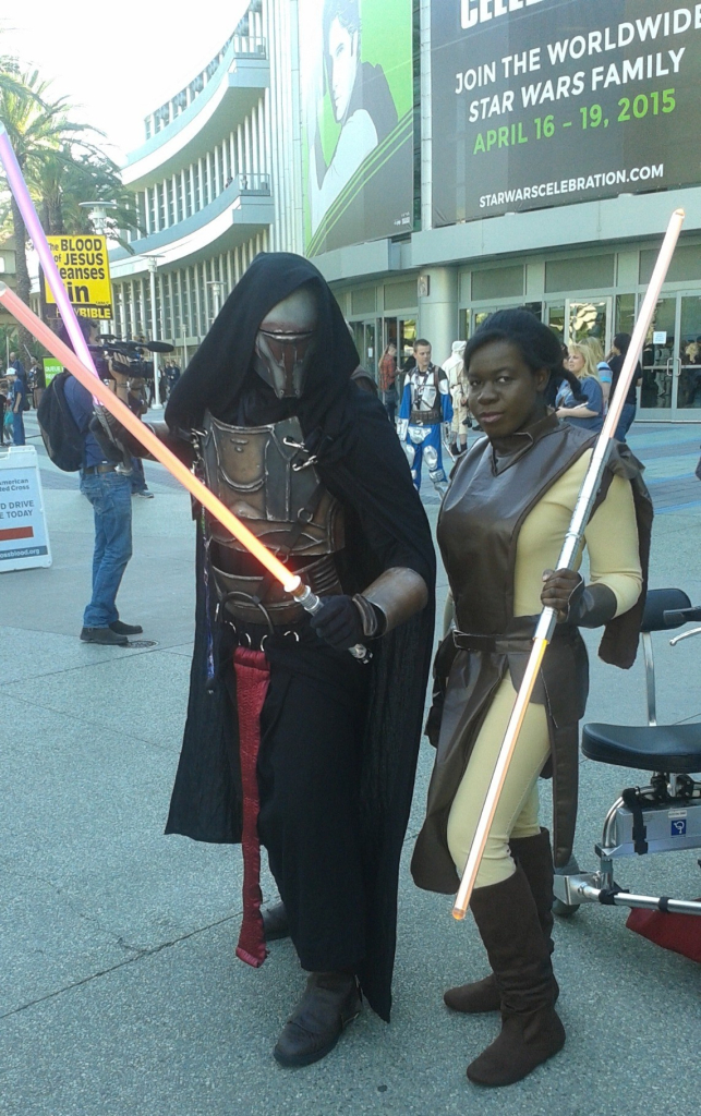 a photo of 2 cosplayers holding lightsabers in front of a convention center during Star Wars Celebration. On the left, one is wearing a black hooded cloak over a helmet that completely covers their face, and matching ornate armor over dark clothing, dressed as Revan, and on the right, the other has dark brown skin, black hair styled in an updo, and is wearing a long brown tabard over a tan bodysuit, with a matching belt and boots. Next to her is her wheelchair.
