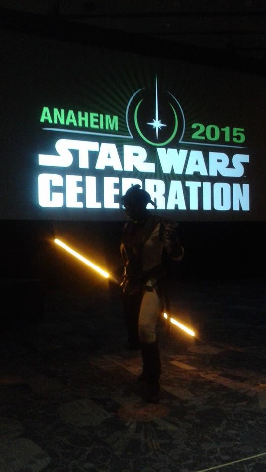 A dark convention room with the only illumination coming from the projection screen which reads 'Anaheim 2015 Star Wars Celebration' and the dimly lit figure in the center, holding a bright yellow double bladed light saber at an angle behind her back. Her face is obscured, but from her silhouette you can tell her hair and clothing are styled to resemble Bastila Shan from Knights of the Old Republic