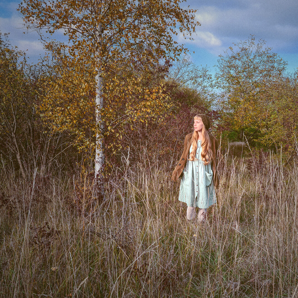 me: lady with long blonde hair in a mint color dress standing in tall grasses with trees