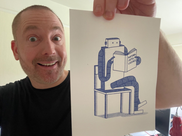 Yours truly (average white-passing guy with short dark brown hair in a black t-shirt) holding up a small blue ink on white paper sketch of a robot sitting in a chair reading a book titled Natural Intelligence.
