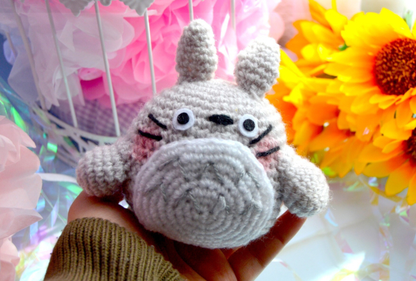 Photograph of a crocheted Totoro soft toy, with flowers in the background.