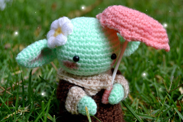 Photograph of a crocheted Yoda soft toy carrying an umbrella and with a flower behind the ear. There is grass in the background.