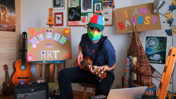 A very silly and chaotic photo of myself sitting in front of the camera playing a bass ukulele. I'm in a silly propeller hat, Hacktoberfest t-shirt, wearing rainbow cloth mask and shiny round sunglasses full of reddish sequins that together cover up the whole face. In the background there's a sign stating that "Games = art" with a rainbow, googly eyes and colorful flowers around. Another sign states "I am Banksy" in big colorful letters. An ukulele and classical guitar are standing in the background, so does a poster for the game "Karambola". Popular "Minions" characters are climbing the wall behind. A wicked wicker "bird" sculpture is standing in the foreground on a djembe. There's also a guitar amp with crocheted small owls standing on top (one in a cardboard car), a paper magazine about a rock band "Perfect", and a Dell laptop with a Librem 5 phone placed below its keyboard, displaying phosh's lockscreen. Color saturation has been visibly boosted.