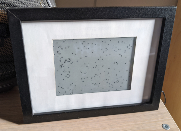 E-ink screen in a frame, with a Conway's Game of Life grid on it. There is a cluster of activity happening on the left side of the grid, representing an ongoing bot attack.