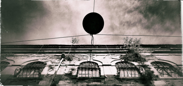 A random street in Istanbul, Turkey. The viewer is looking straight up into the sky. An extinguished streetlight is hanging directly overhead. A building stretches across the bottom half of the view. The building looks old and careworn. Plants are growing out of the walls. Delicate, black wrought-iron bars cover the windows.