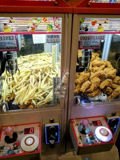A photo of a two amusement arcade claw machines. The one on the left has fries in it. The one on the right, fried chicken.