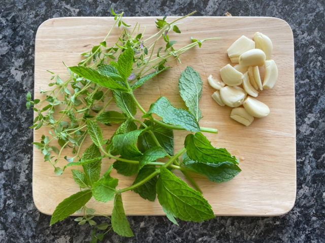 Photo of fresh thyme, mint, and garlic on a small wooden board on a dark kitchen counter.