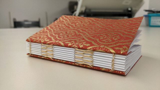 small paperbound book demonstrating a sewing method for a bookbinding class. Colors are red and gold.