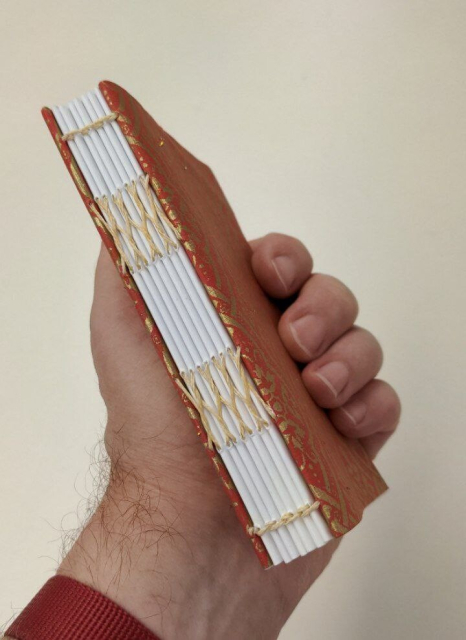 small paperbound book held in a man's hand, demonstrating a sewing method for a bookbinding class. Colors are red and gold.