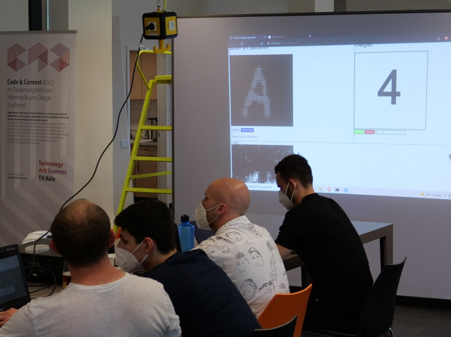 4 students sitting in front of a screen that displays a tensorflowjs perceptron-model developed by students. The model has been trained to recognize numbers on the MNIST Dataset, here being tested on new data recognizing an A as a 4, the interface allows online re-training.