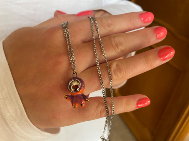 Close-up of a little orange and purple ceramic astronaut pendant with metallic face plate on a thin silver chain bring demonstrated by a female hand with orange nails 