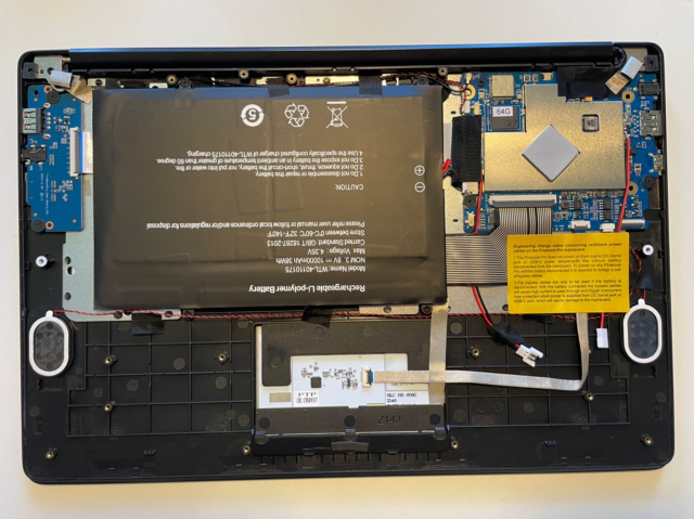 Photo of inside of a PineBook Pro. A large battery takes up most of the space with a small motherboard on the top-right and what looks like an I/O board with USB, line, and SD card in on the top-left. Also visible are speaker grills and a bypass cable with a note about its use.