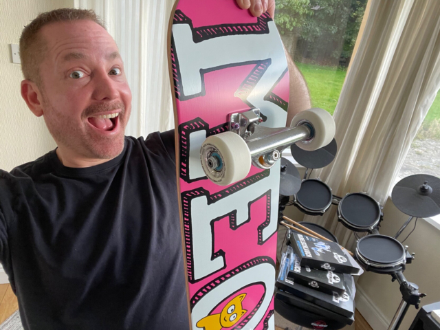 Me holding up a skateboard with meow written in large letters on a pink background with a yellow kitten looking out from the “o”.