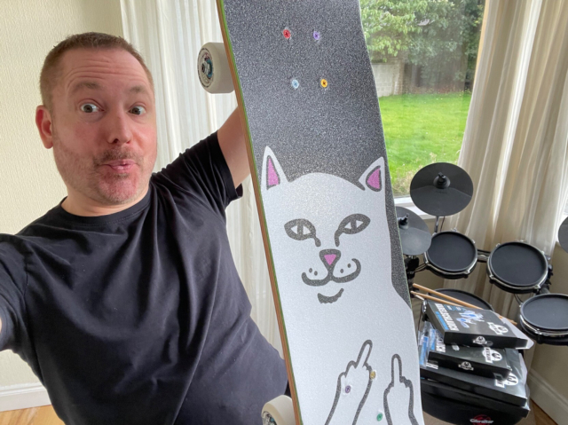 Me holding up a skateboard with the graphic of a white cat giving both middle fingers on the grip tape.