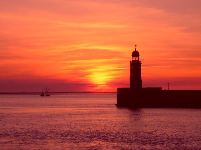 A waterscape photo depicting the sharp black silhouette of a lighthouse against the backdrop of a deeply orange and yellow sunset over the water. Photo taken at the harbor in Bremerhaven, northern Germany. 