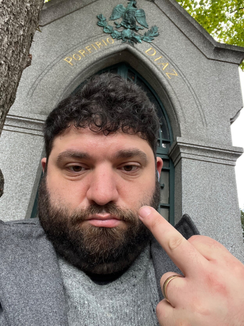 A large bearded man facing the camera makes a rude gesture, behind him is a grave marker that says “Porfirio Diaz” in gold letters. It has the eschucho of Mexico on it