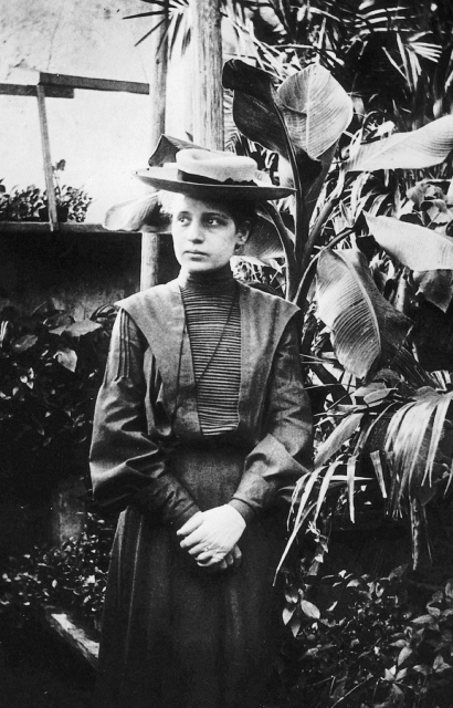 Black and white photo of Lise Meitner. She is standing in what appears to be a greenhouse, as there are several large plants behind her. Meitner is wearing a dark dress and blouse, with a high collar and hat. She is looking to the left of the photographer, with her hands clasped in front of her.