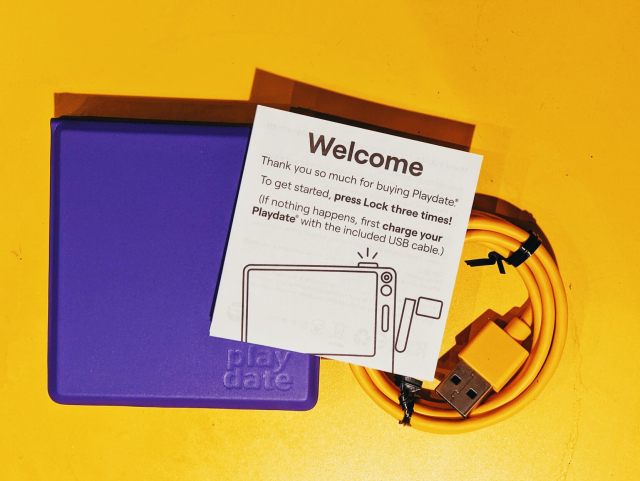 A Playdate console in a purple cover with the instruction leaflet and the yellow charging cable on a yellow background.