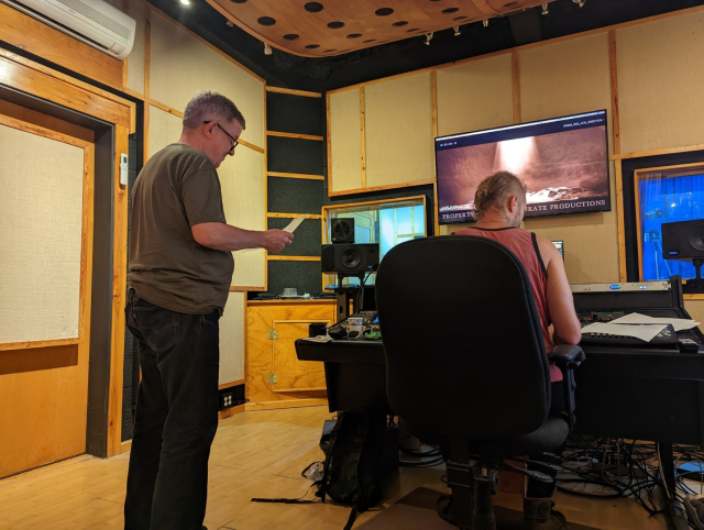 Douglas Mackinnon on the left stands looking at a script and listening. He is in an ADR studio in Kingston. The engineer has his back to us in front of the control desk.