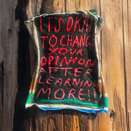 Homemade patch with worn edges nailed to a wooden post. Bright red, handwritten capital letters read: "It's okay to change your opinion after learning more!"