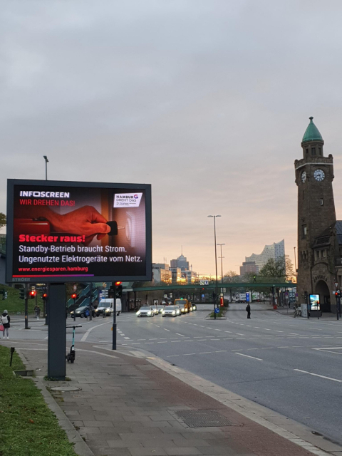 Hamburg, Landungsbrücken. In der Ferne die Elbphilharmonie. Eine breite Straße, wenig los. Neben dem Turm eine grelle Werbetafel. Auf dem Gehweg vorne eine zweite, riesige digitale Werbetafel mit dem Motiv "Hamburg dreht das. INFOSCREEN wir drehen das! Stecker raus! Standby-Betrieb braucht Strom. Ungenutzte Elektrogeräte vom Netz."