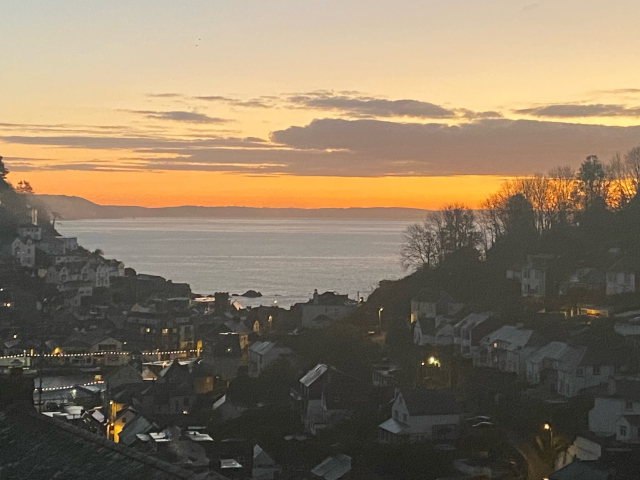Bedroom window view looking at the sea at sunrise. The sky is slightly orange and a few clouds are hovering over the sea. Surrounded by old cottages in a town in Cornwall, Southwest England. 