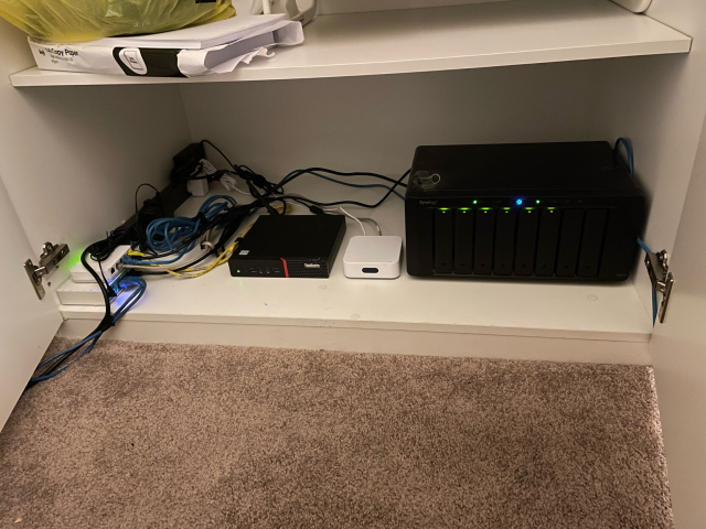 A picture of hailey’s home datacenter. A bundle of cables (power and ethernet) leads into a cupboard containing a router, extra switch, little thinkcenter, WAP, and synology nas