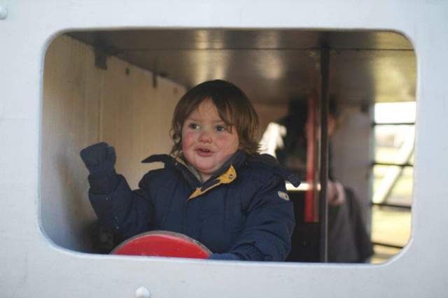 Small boy driving a wooden bus in a playground, we're looking through the (nonexistent) windscreen at him holding the red wooden wheel with one hand and waving his other arm around.