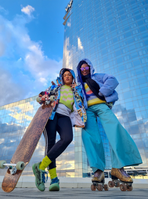 Two femmes are standing front large glass buildings.  The sky and clouds are reflected. 
The one on left is wearing pink and yellow sweater and multicolor jacket. They have a longboard with plant illustrations 


Femme on right is wearing a multicolores striped sweater with a sky blue puffy coat and blue wide leg pants. She has cheetah print quad roller skates