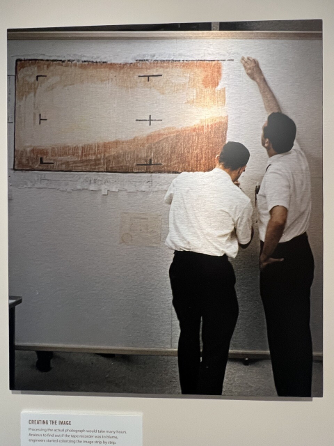 Historical picture of two men, each wearing indentical white dress short-sleeved shorts, black pants, black belts, and having short black cropped hair standing in front of the pastel Mariner map on a white drywall wall at NASA/JPL. Image from July 14, 1965. Official caption reads, “Telecommunications engineers at the Jet Propulsion Laboratory putting the finishing touches on their color-by-number version of Mariner 4's digital data.”