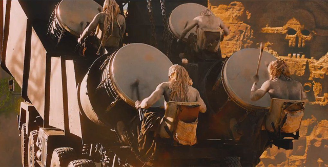 Picture of an escene from Mad Max, showing four giant drums mounted on a large vehicle, beaten by four rude guys.