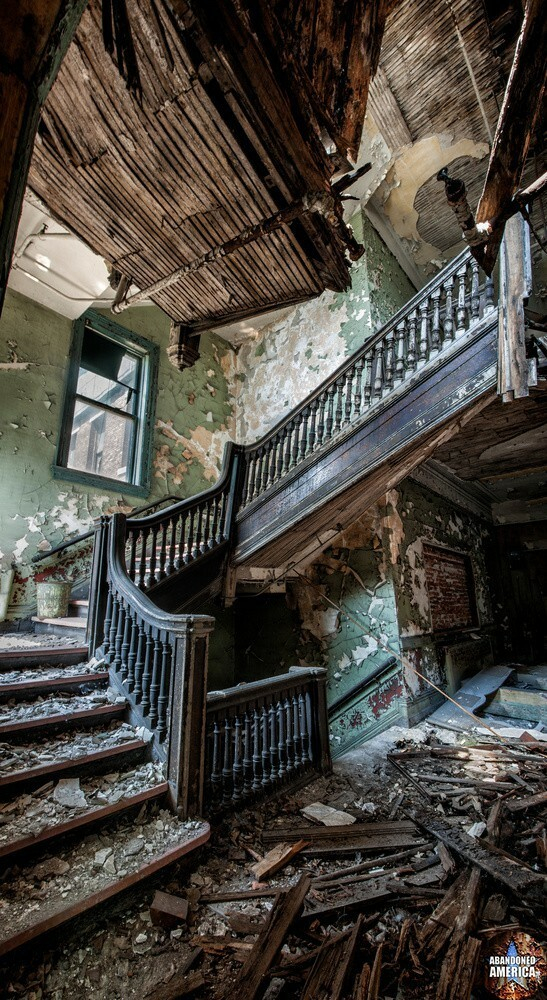 A view up a crooked wooden staircase. The walls are a mint green that is peeling away to reveal white underneath. Another stairwell leading down is mostly obscured. High above the first landing there is a window and a pail sits on one of the steps. To the right another hallway leads off into darkness.