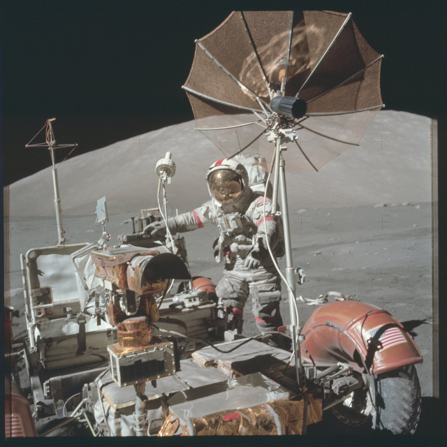 One of the Apollo 17 astronauts standing with the rose. Their visor is a reflective gold color, while copper-colored foil wraps many of the components of the rover. A large, umbrella-like radio dish is mounted on the from of the rover. A large gray hill rises in the background.