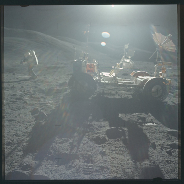A color photo of the Apollo 17 lunar rover. The sun is just outside the frame, causing long shadows and substantial optical artifacts. An astronaut in the distance is seen in profile. The rover has a large radio dish mounted on the back.