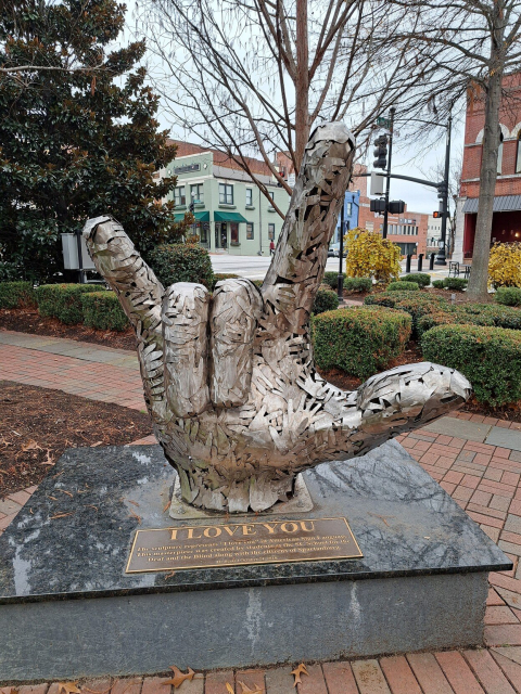 Metal sculpture of a hand signing I Love You in ASL. It is made of smaller hands, some open and some also making the same sign. In the background are trees, bushes, buildings, and an overcast sky.