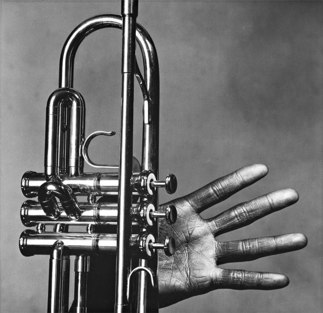 Black and White photo of Miles Davis' left hand against a murky gray background, fingers splayed around a trumpet