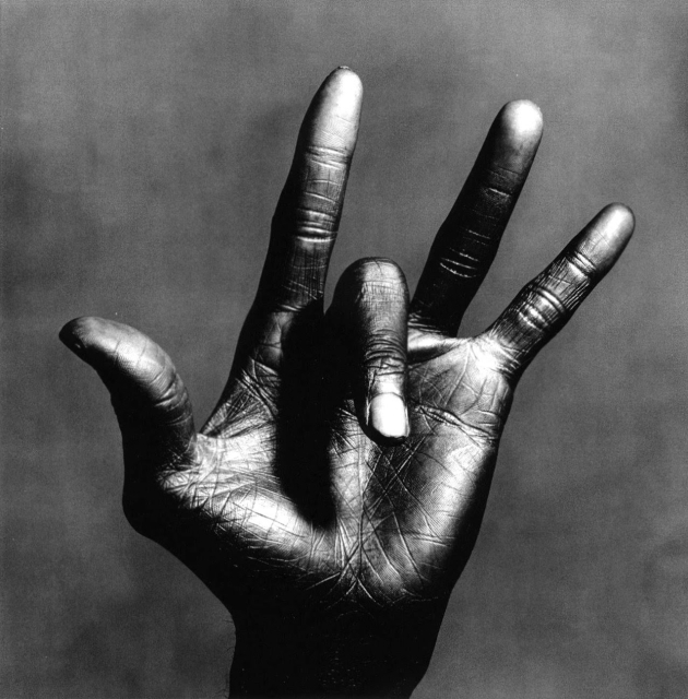 Black and White photo of Miles Davis' left hand against a murky gray background, thumb hooked back, middle finger folded over, other fingers splayed