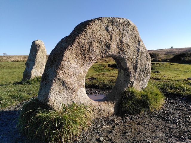 Photo of Men-an-tol in Cornwall. A stone circle with a normal standing stone behind it, in the sun, with a mine in the far distance 