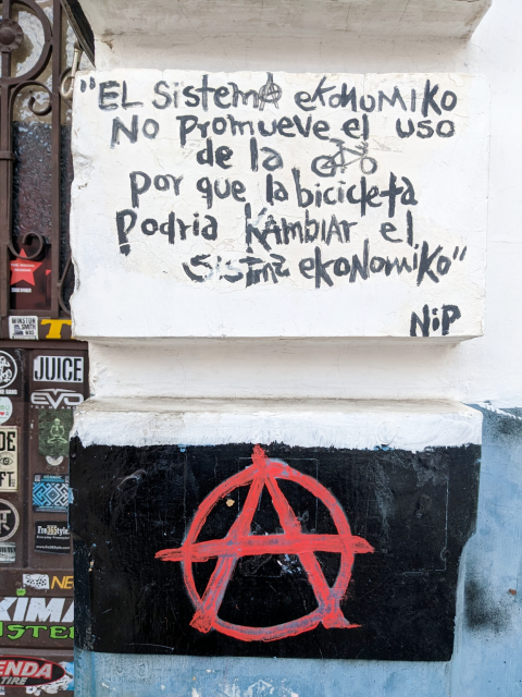 Advertencia en el muro afuera de una tienda de alquiler bicis en San Cristóbal de las Casas, Chiapas que dice "El sistema economico no promueve el uso de la bici por que la bicicleta pordria cambiar el sistema economico" con el simbolo anarchista en rojo y negro.