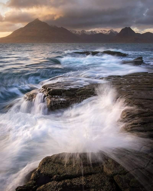 Waves crashing over rocks, a wild sea and the cullin mountains in the distance at sunset