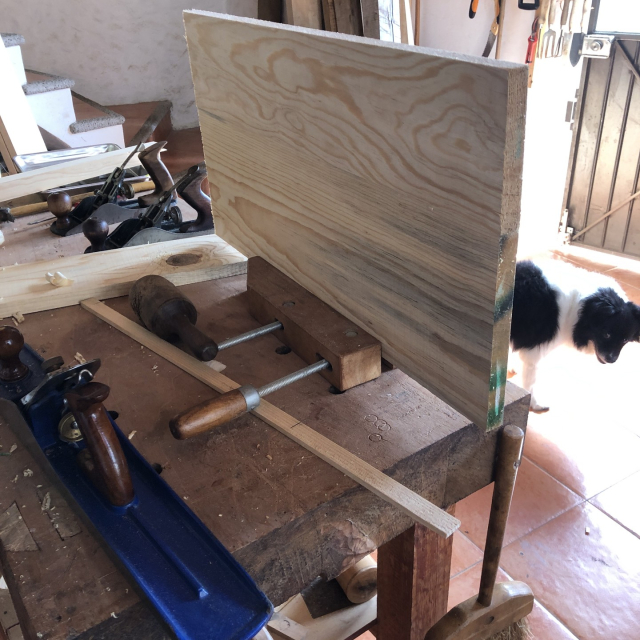 Sargento de madera puesto sobre el banco de trabajo, sin apretar, para nada más sostener las tablas en lo que se verifican. Mozzarello explora el taller por ahí al fondo.