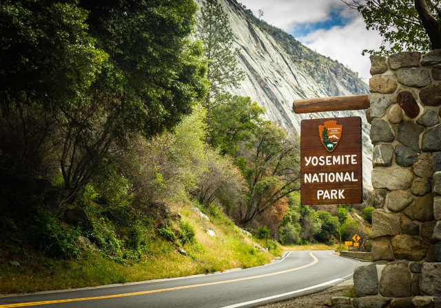 Picture of the entrance to the Yosemite National Park with a plaque saying Yosemite National Park hanging from a pile of rocks on the side of the road.