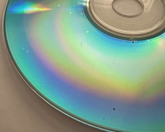 the underside of a CD-R, a vibrant blue color with rainbow arcs due to thin film interference. there are several small spots visible, with a brighter outer ring enclosing a variable sized dark center where the top disc layer has eaten away