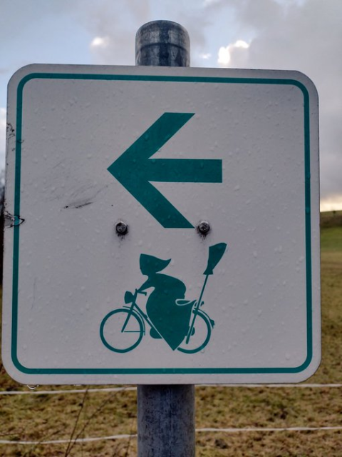 a green & white cycling route direction sign, the arrow points to the left. bottom half of the sign is a green icon of a witch cycling a bike, with a broom stashed on the back.