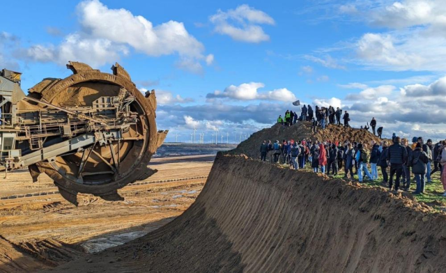 Climate activists standing against the inhuman-scale machinery of the open-pit Lützerath brown coal mine, with a wind farm visible in the distance.