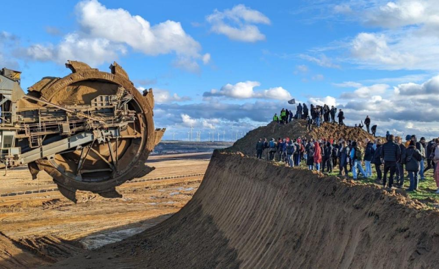 Climate activists standing against the inhuman-scale machinery of the open-pit Lützerath brown coal mine, with a wind farm visible in the distance.