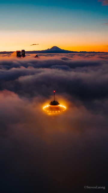The Space Needle rises above the fog at dawn in Seattle, Washington. A couple of skyscrapers and Mt Rainier are visible in the distance