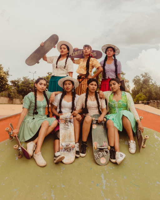 An empowering photo of seven Indigenous Bolivian women posing at a skatepark. Wearing traditional garments, they hold their skateboards proudly and look to the camera.